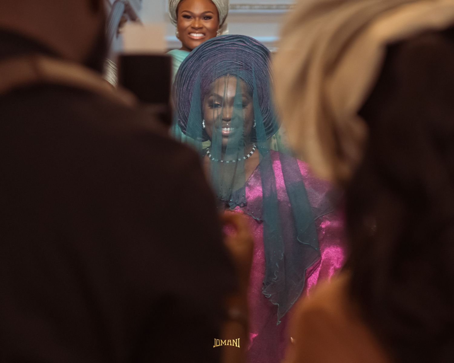 Woman in pink outfit and blue traditional Nigerian gele headwrap smiles at a festive celebration.