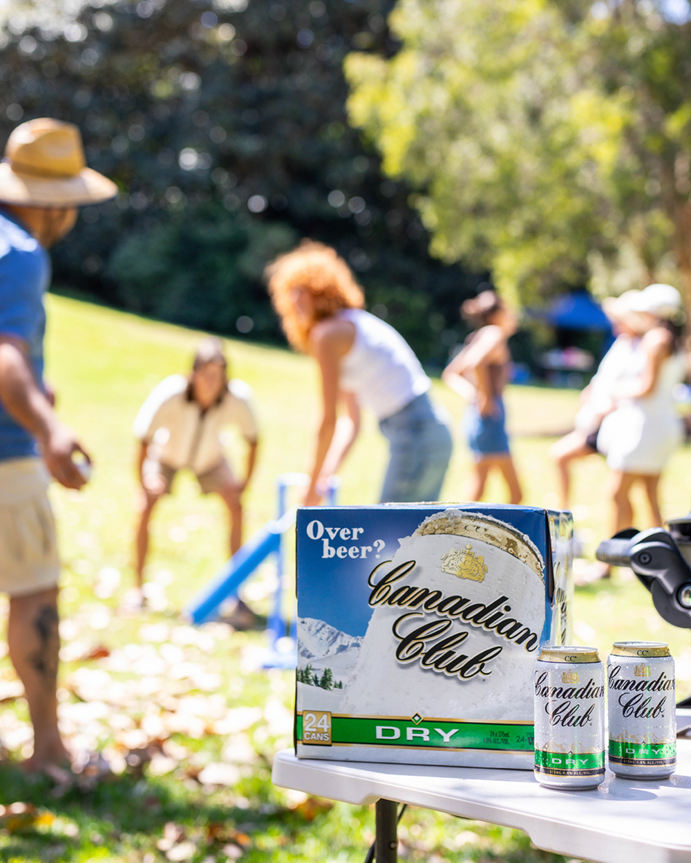 Canadian Club Dry beer cans on table with friends playing lawn games in sunny park.