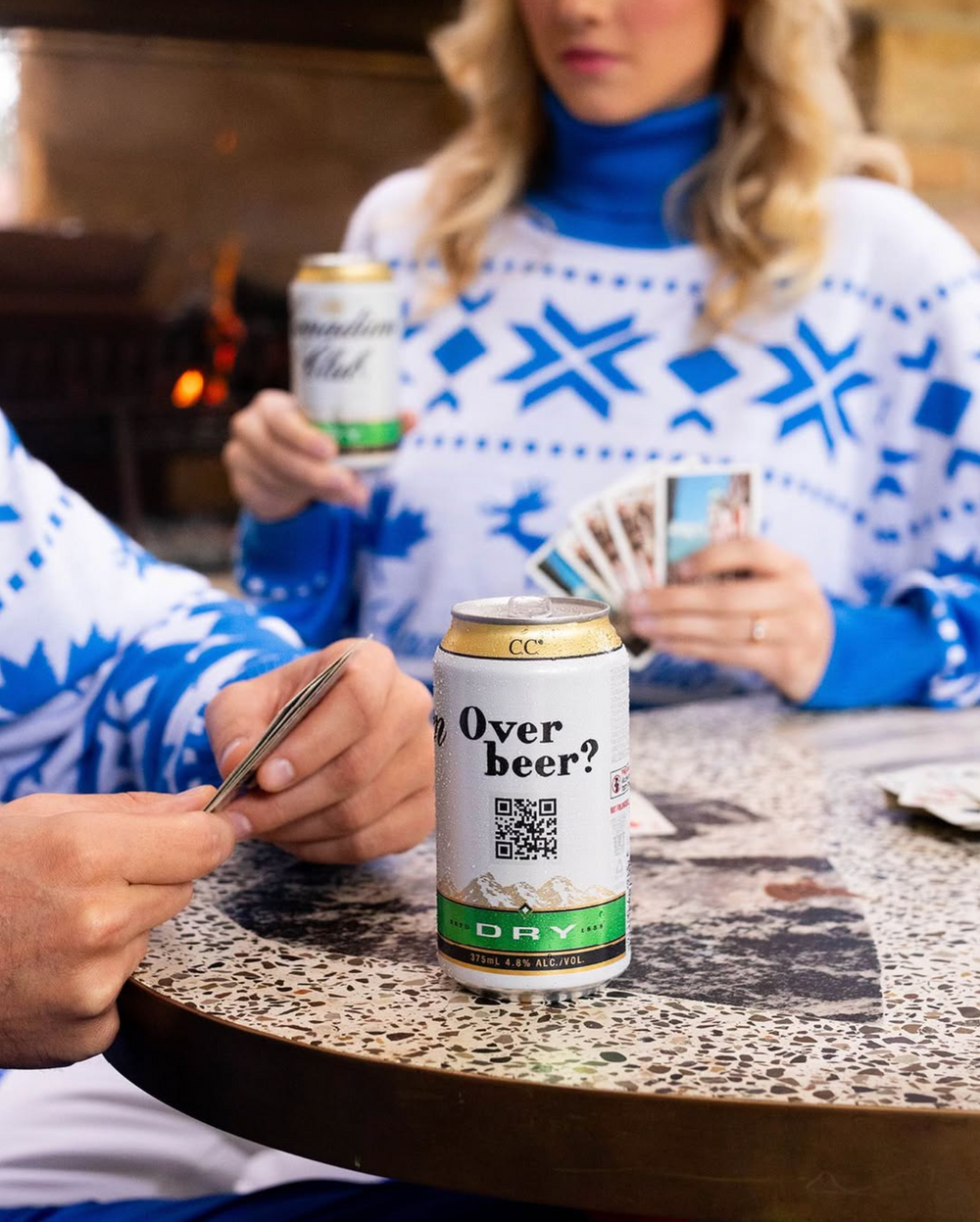 Couple in matching blue Christmas sweaters playing cards with Canadian Club Dry beer cans.