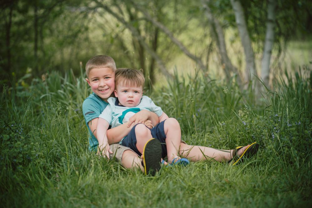 Two children sitting together in tall green grass during a summer photoshoot.