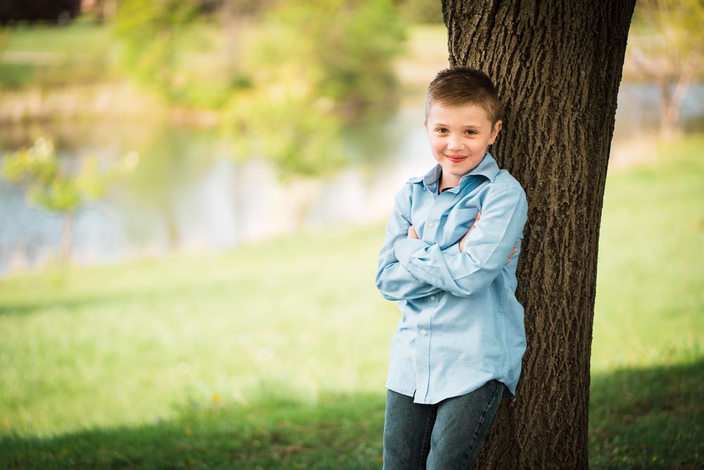 A young person in a light blue shirt leans against a tree trunk in a park setting.