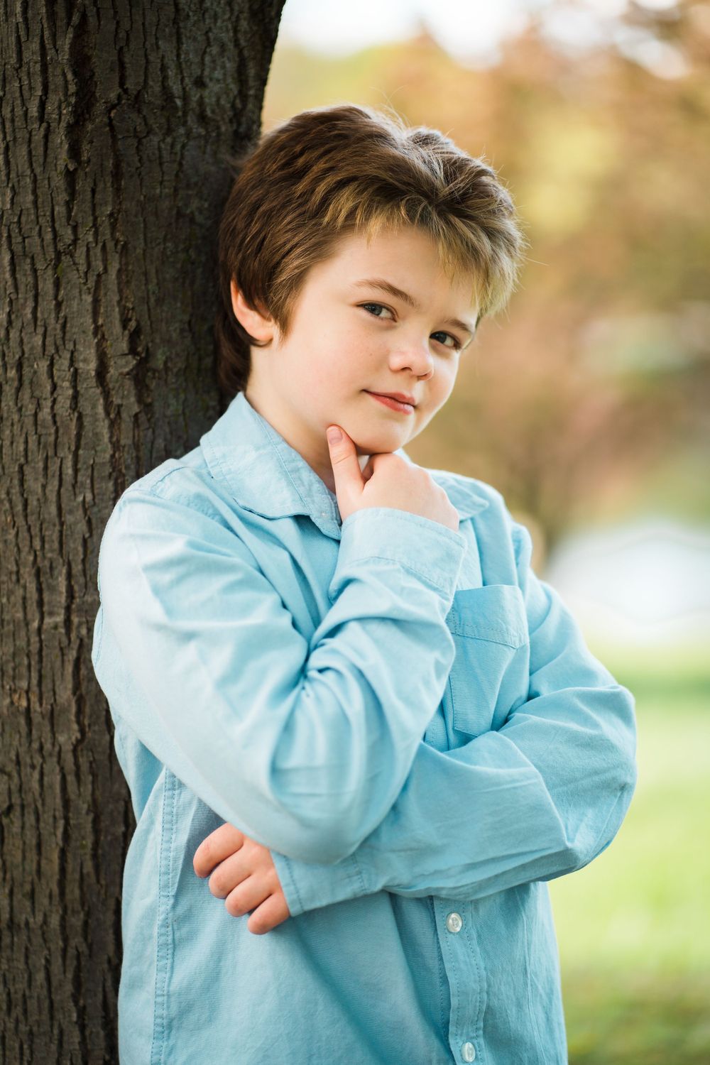 A youth in a light blue button-down shirt poses thoughtfully against a tree trunk.