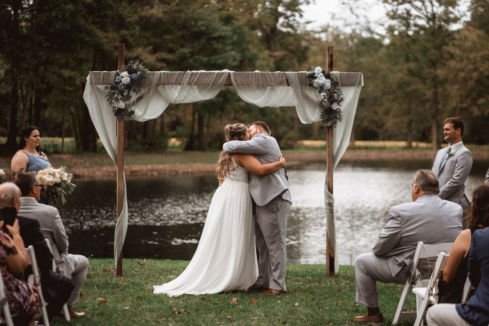 Wedding ceremony by a lake with guests watching the couple embrace under a decorated arch.