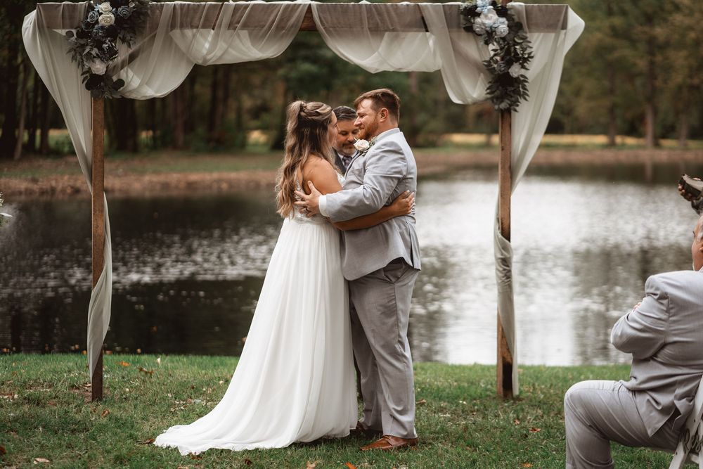 A couple shares their first kiss under a floral arch during an outdoor lakeside wedding ceremony.