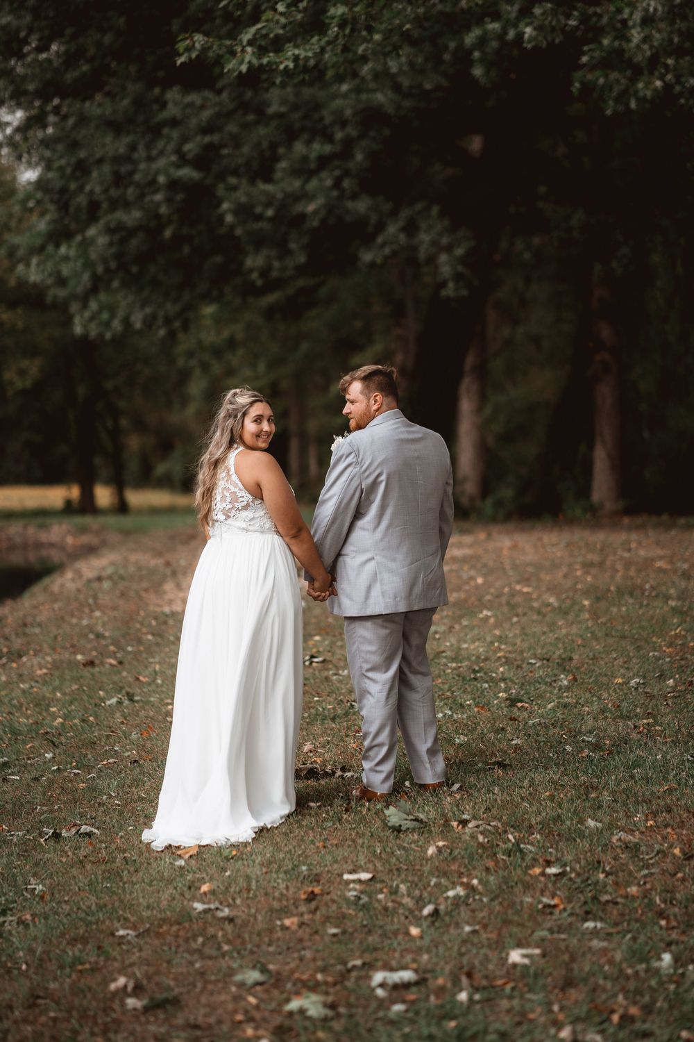 A couple in wedding attire holds hands while walking through a wooded area.