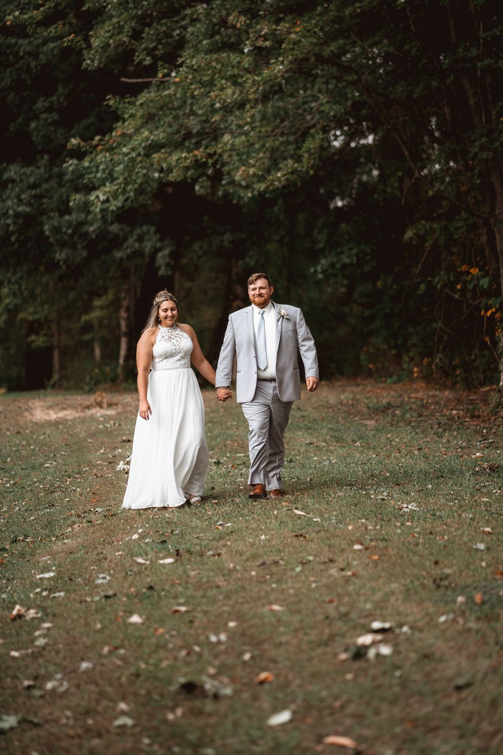 Newlyweds walk hand in hand on a path surrounded by autumn trees.