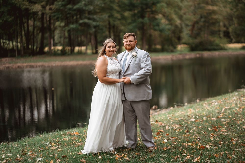A couple poses for their wedding portrait by a tranquil lake surrounded by fall foliage.
