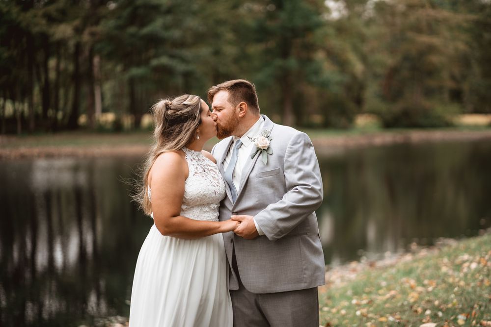 A tender wedding moment captured by a lakeside with fall foliage in the background.