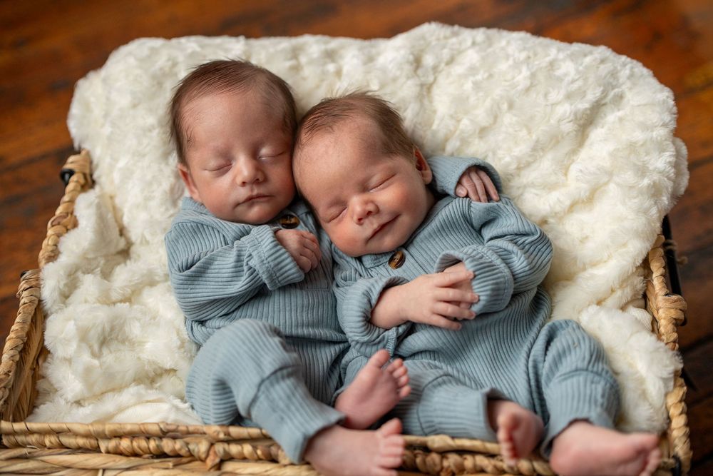 Sleeping newborn twins dressed in matching blue outfits nestled together on a cream-colored blanket.