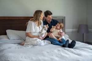 A family sits together on a white bed while looking at a mobile device.