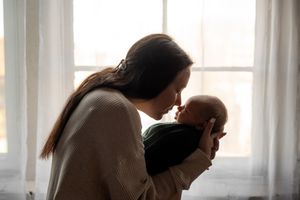 A tender moment between parent and baby is captured in silhouette by a sunlit window with sheer curtains.