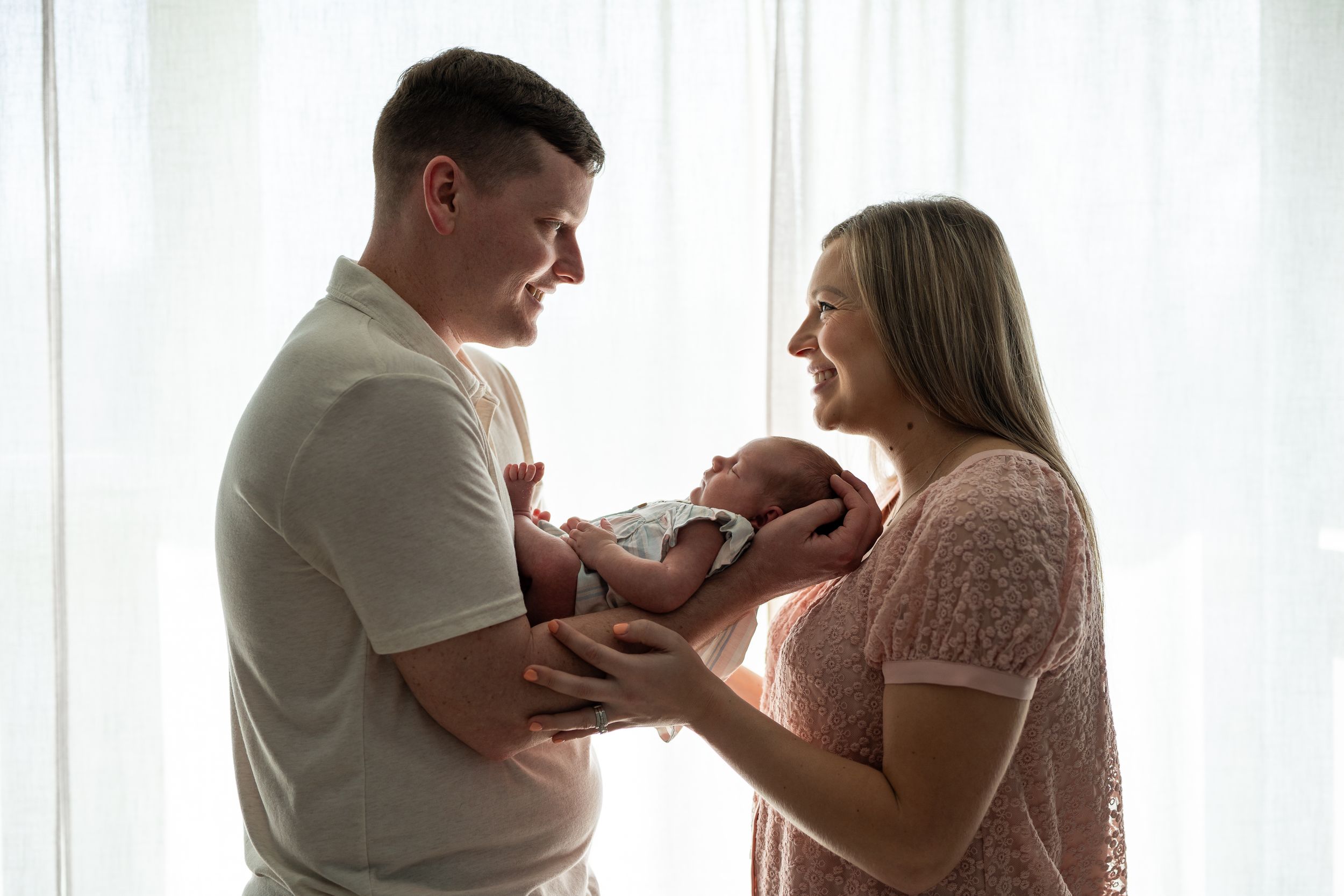 A tender newborn photography session shows intimate family moments by a bright window in soft natural light.