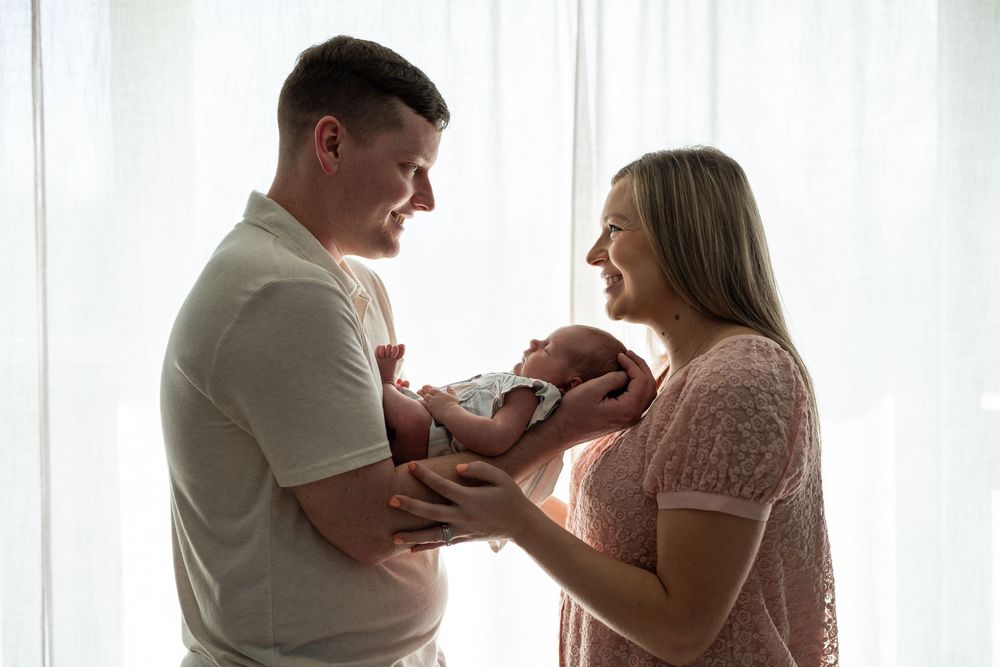 A tender newborn photography session shows intimate family moments by a bright window in soft natural light.