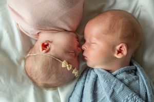 Two sleeping infants lying face to face on white bedding, one wearing a blue sweater.