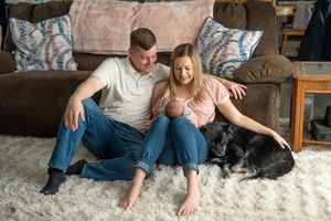A couple relaxes on a plush white rug with their black dog while sitting against a brown couch in a living room.
