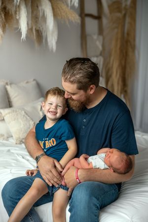 Family sitting together on white bed sharing tender moments with newborn baby in blue room.