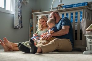 Family sits together on nursery room floor reading a book near a wooden crib.