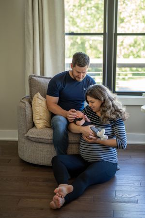 A couple sits together on a floor cushion while looking at a mobile phone in a bright room with hardwood floors.