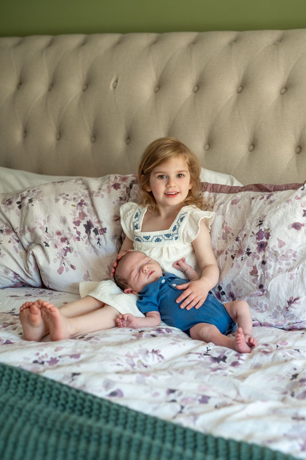 Two children lay together on a floral patterned bed with tufted headboard in soft natural lighting.