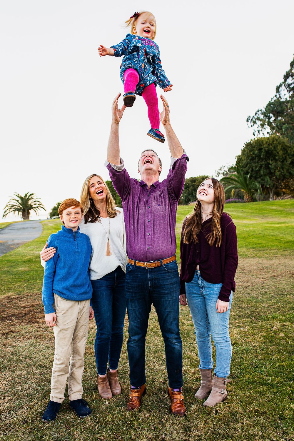 A joyful family of five outdoors, dad tossing laughing toddler girl in the air while mom and two kids watch and smile.