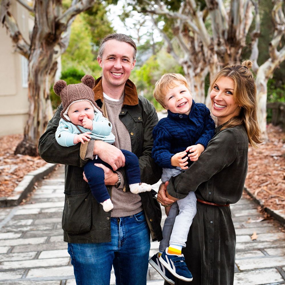 A smiling family of four poses outdoors, with a man and woman each holding a young child, surrounded by trees.