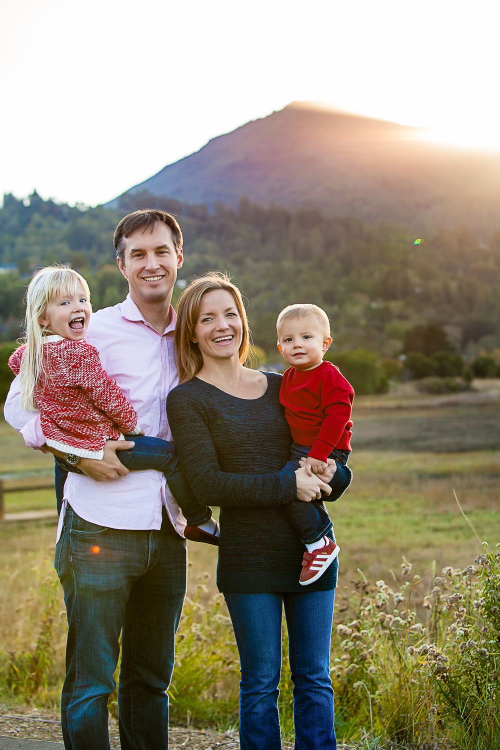 Happy family of four posing outdoors with a sunlit mountain backdrop, parents holding two young children in red outfits.