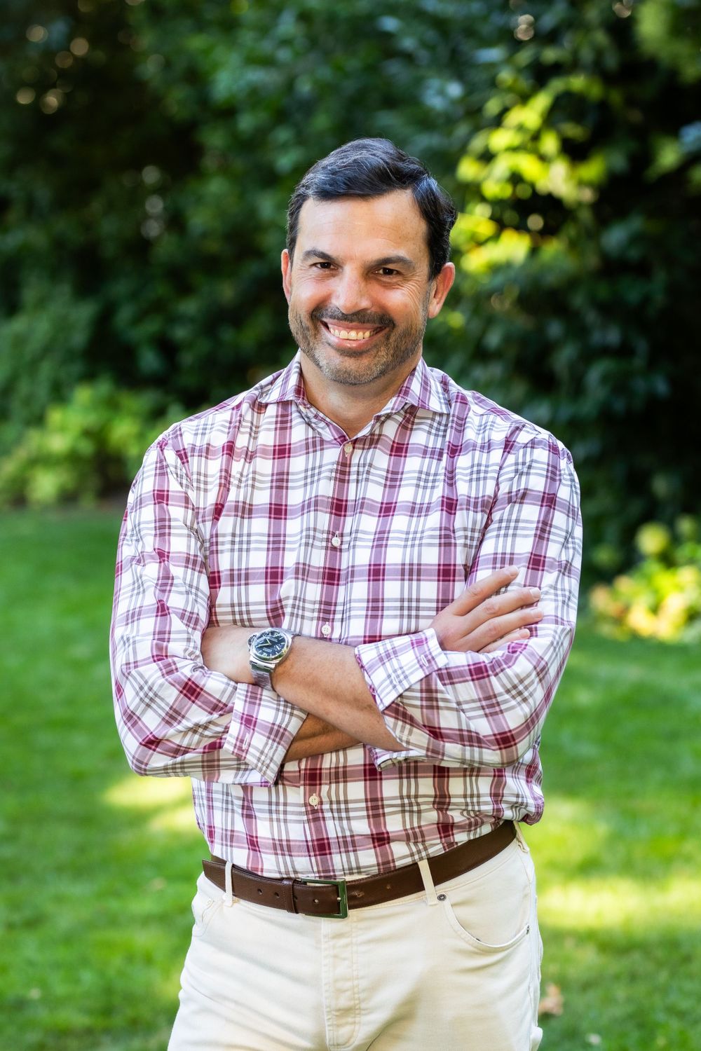 Man wearing plaid button-down shirt smiling outdoors against green foliage background.