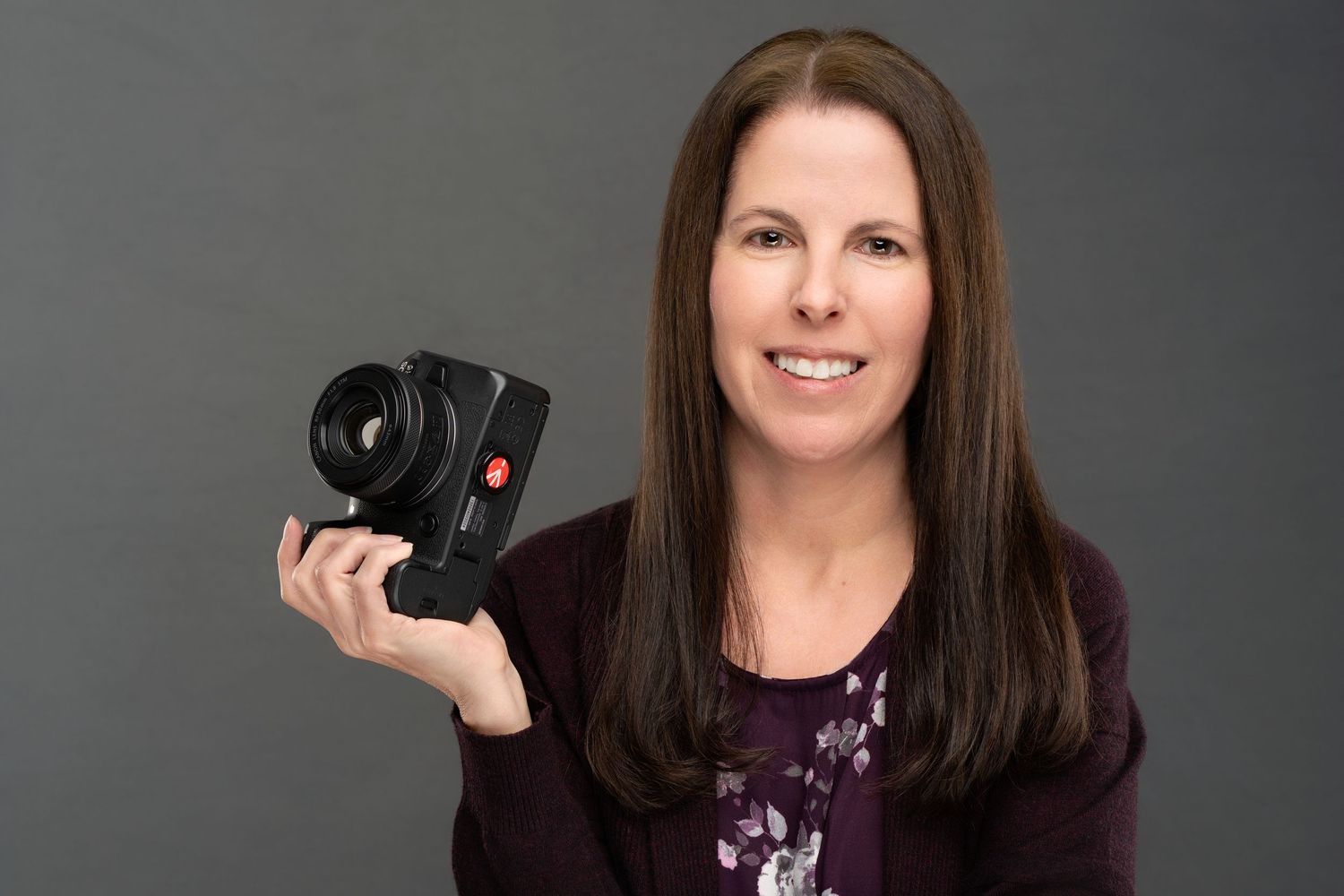 Professional photographer poses with camera equipment against dark grey studio backdrop.