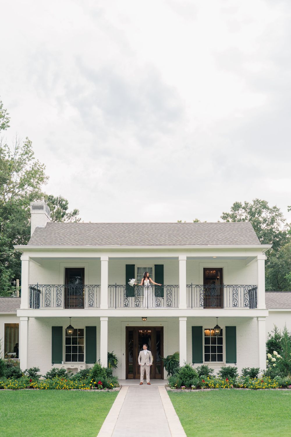 Bride and groom pose outside a grand white Southern plantation-style wedding venue with green shutters and balcony.