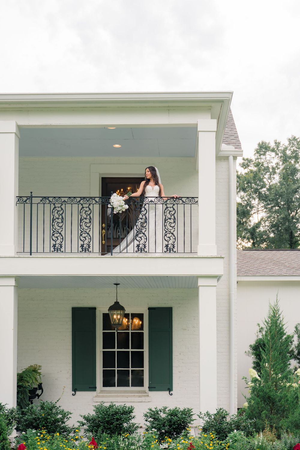 Bride in black and white gown holding bouquet on ornate balcony of elegant white Southern mansion.