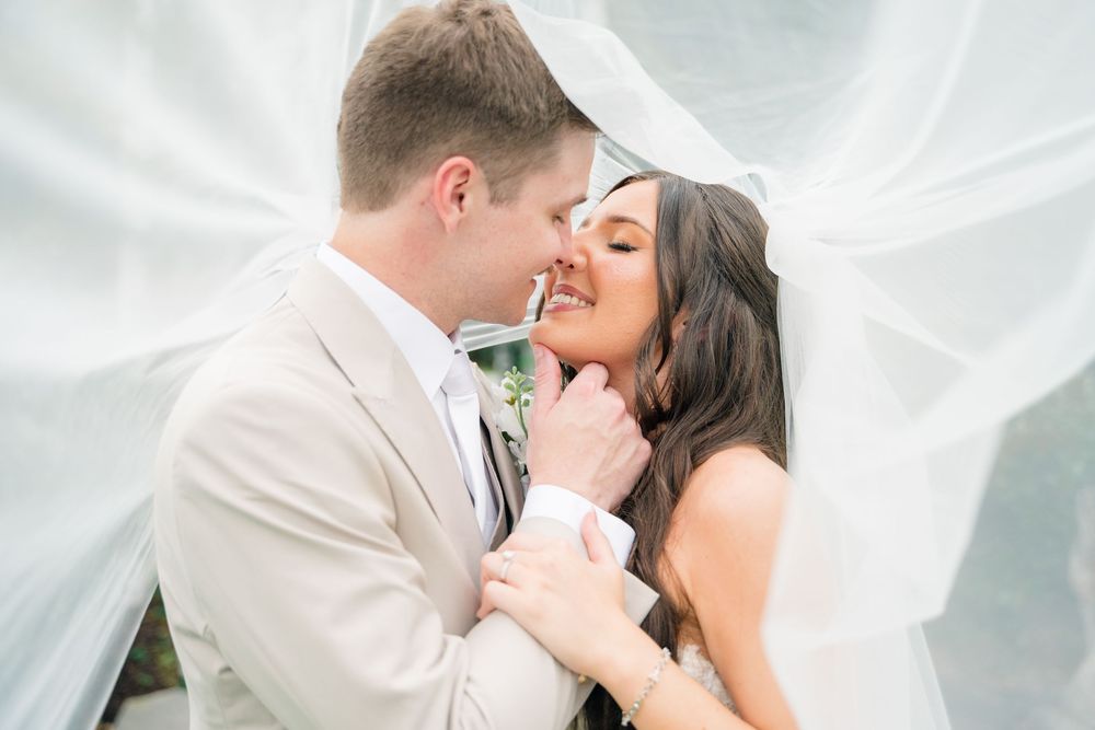 Bride and groom sharing a romantic moment under a flowing white veil, smiling and embracing on their wedding day.