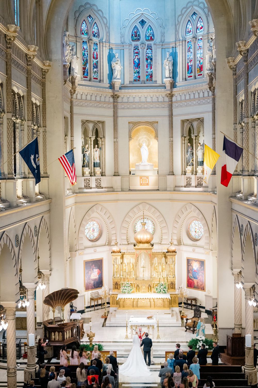 Wedding ceremony inside ornate cathedral with stained glass windows, flags, and golden altar as couple stands at the front.