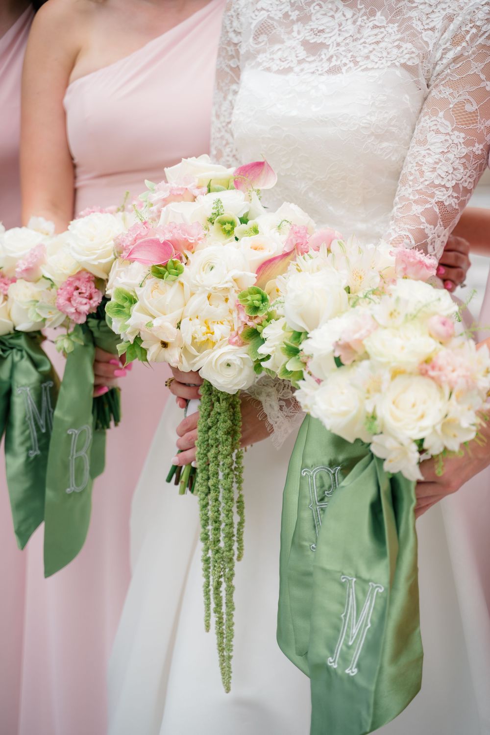 Bride and bridesmaids holding white and pink floral bouquets with green monogrammed satin ribbons at a wedding.