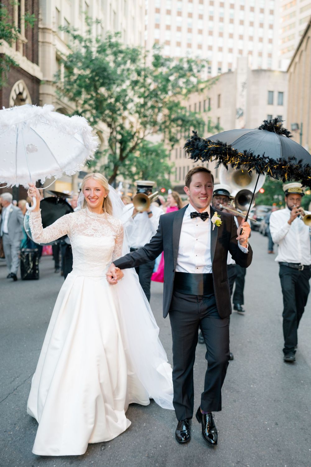 Bride and groom parade through New Orleans streets holding decorative umbrellas with jazz band during second line wedding.