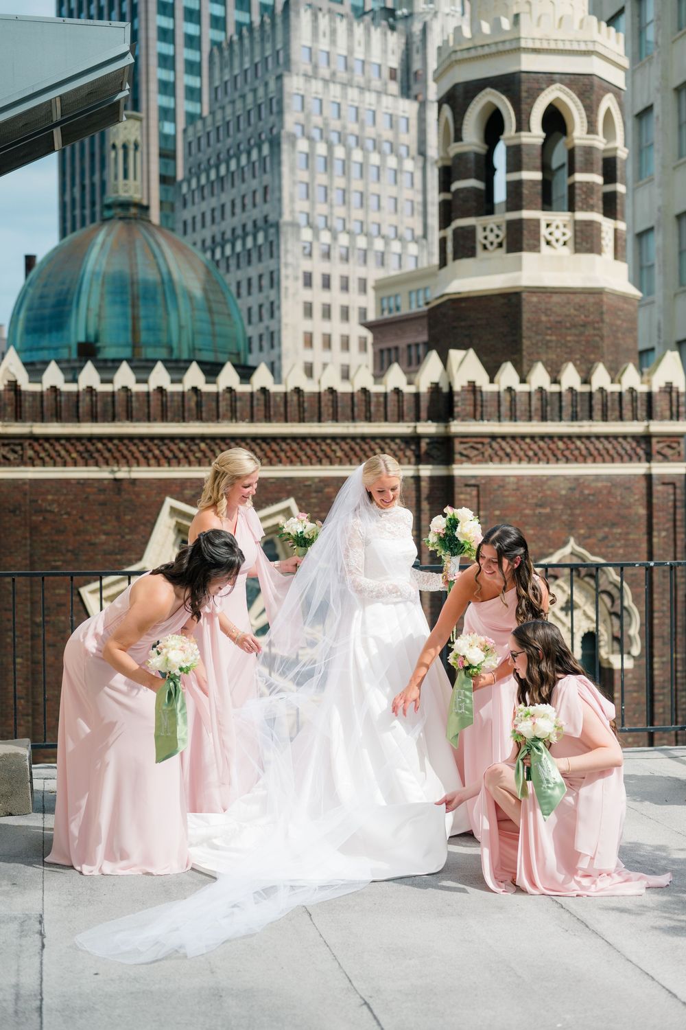 Bride in white gown and veil surrounded by bridesmaids in pink dresses holding bouquets on urban rooftop.