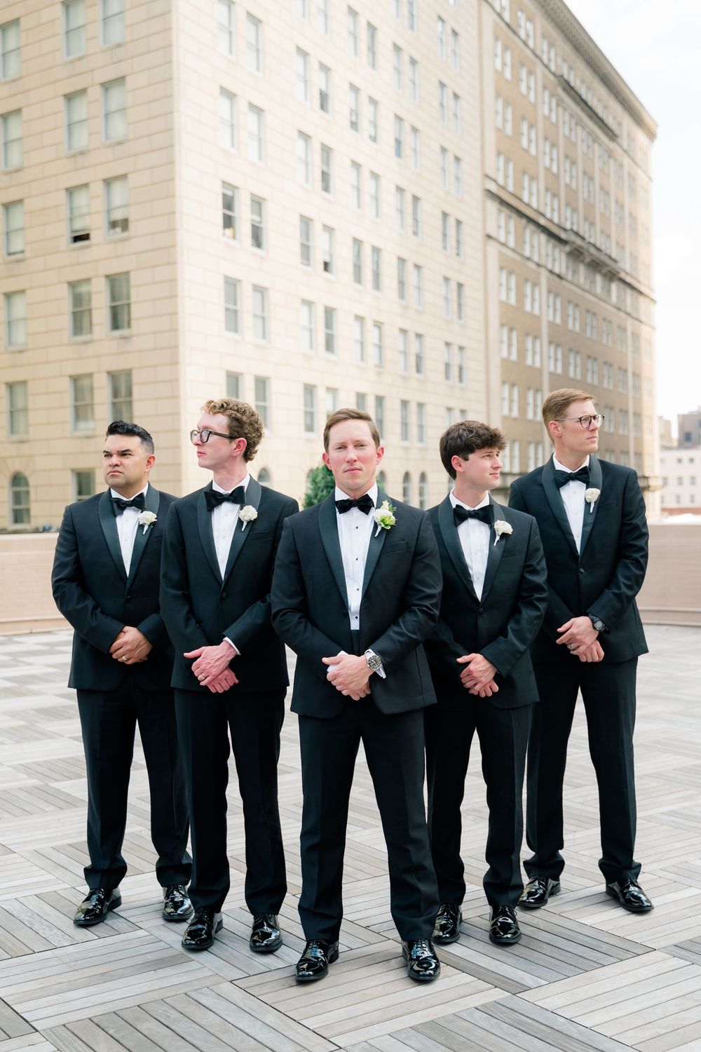 Five groomsmen in black tuxedos with bow ties and boutonnieres pose on a rooftop terrace with a city building backdrop.