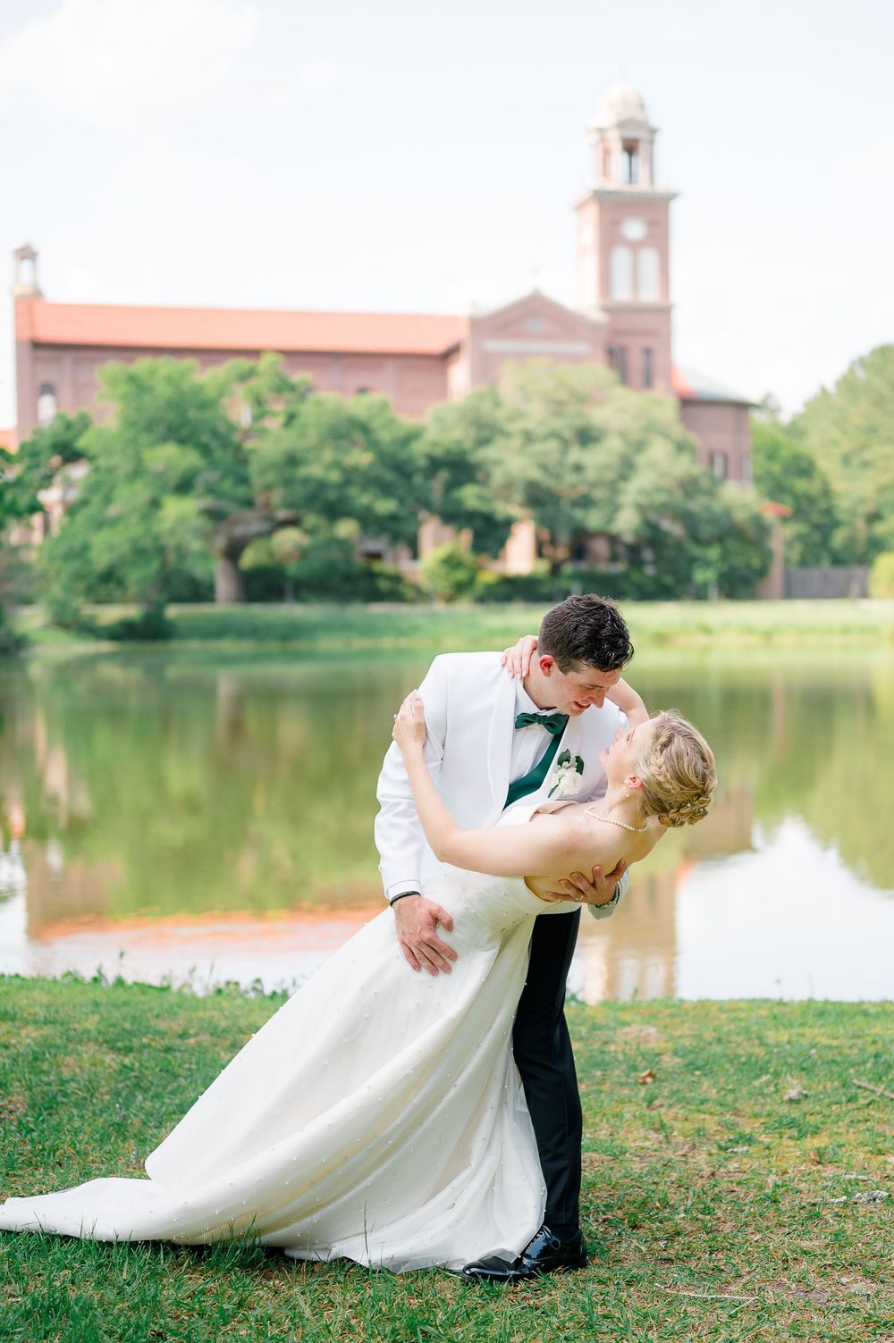Romantic wedding couple shares intimate moment during lakeside photo shoot with historic church building in background.