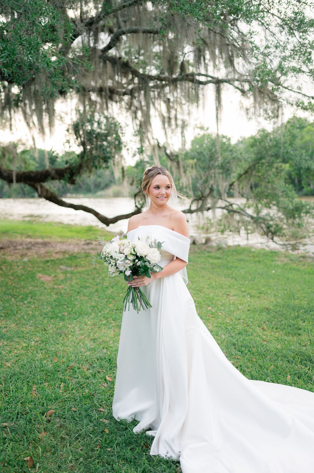 A bride in a flowing white gown stands on a grassy lawn with Spanish moss draped trees in the background.