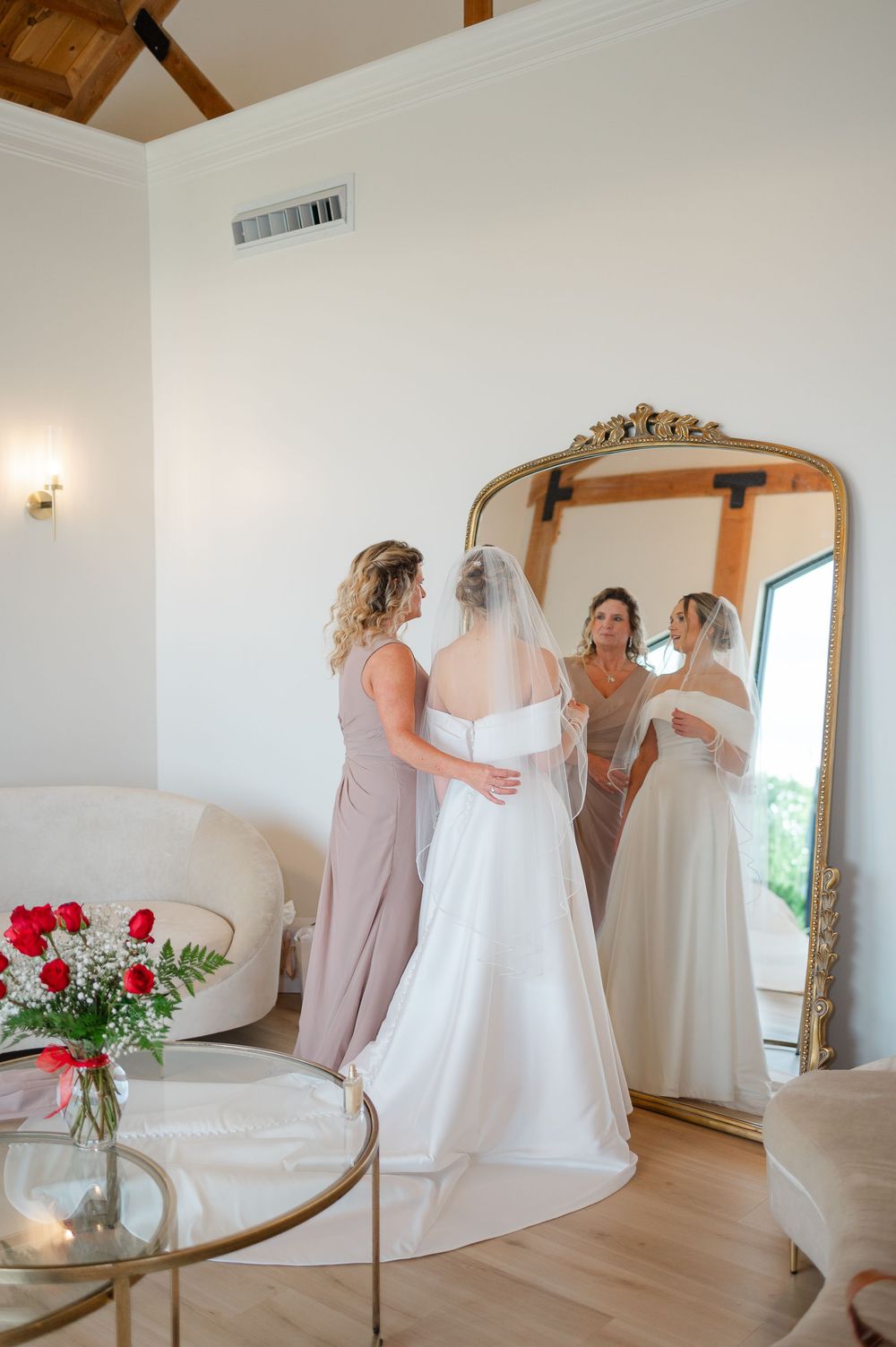 Bridal party in soft pink dresses helps bride prepare in elegant white room with ornate mirror and floral arrangements.