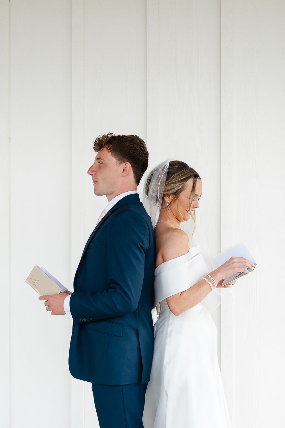 A bride and groom stand back to back reading books against a white wall, she in white dress and he in navy suit.