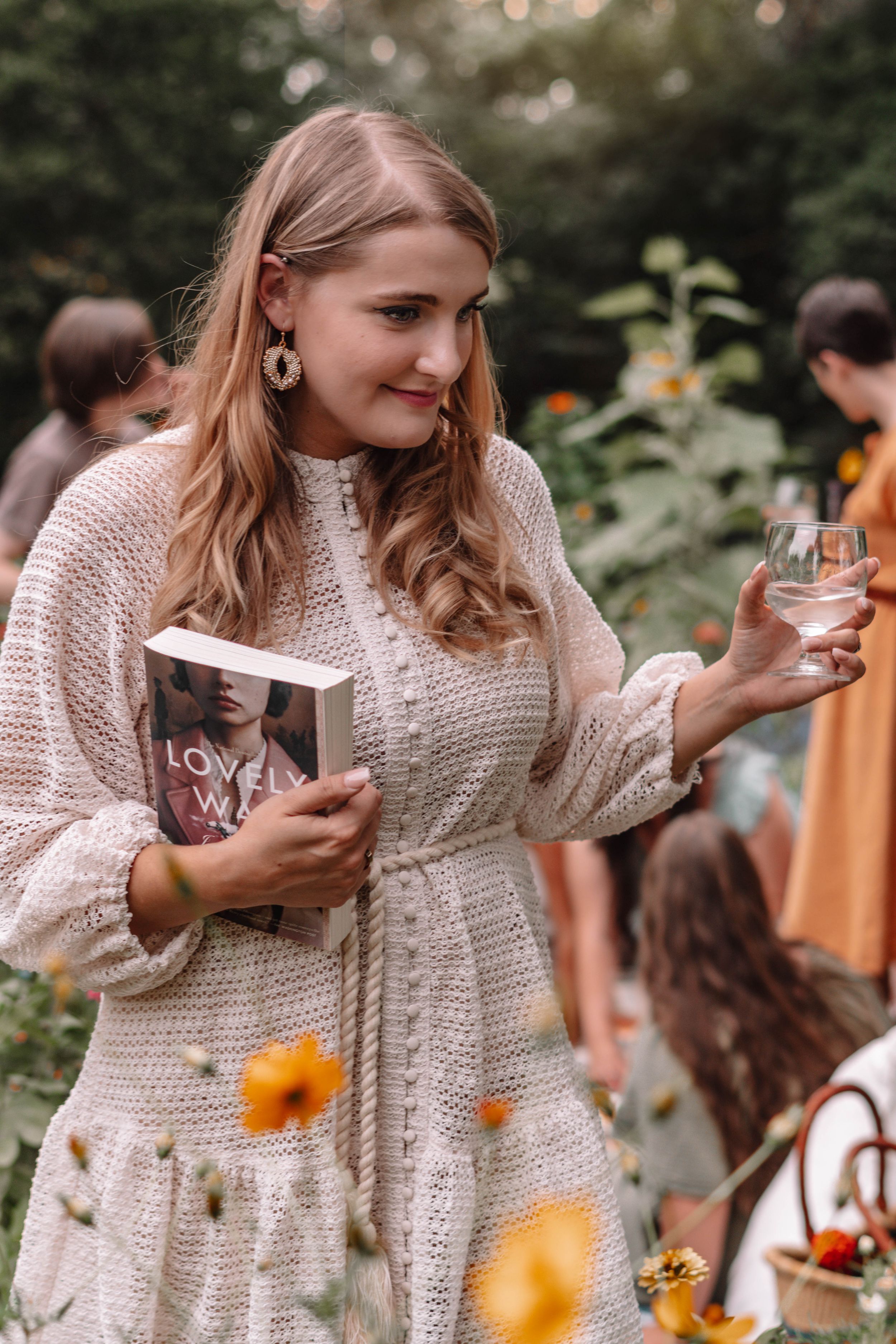 Woman in cream sweater reading intently in a garden with flowers.