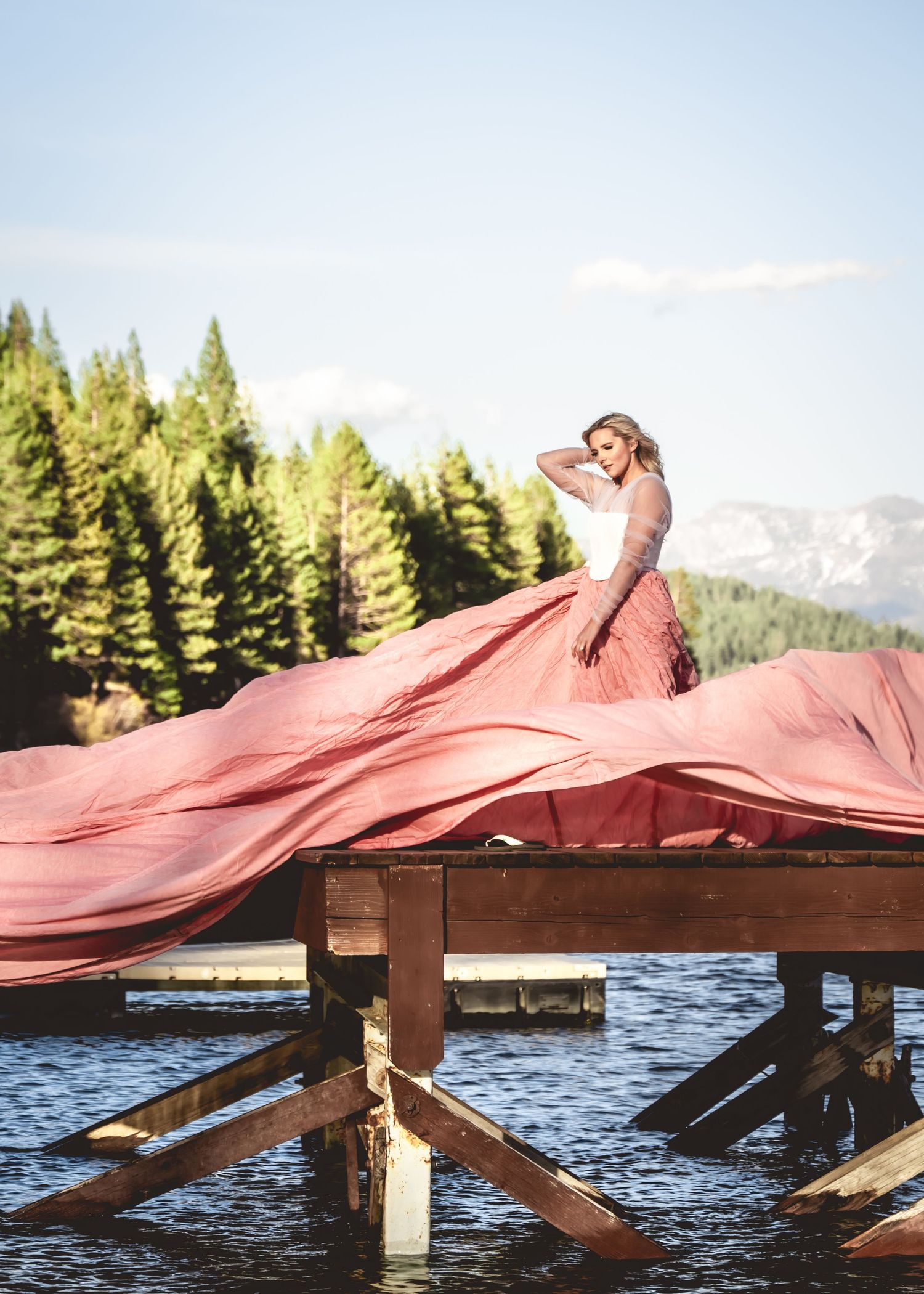 Woman in flowing pink ballgown standing on wooden dock over mountain lake.