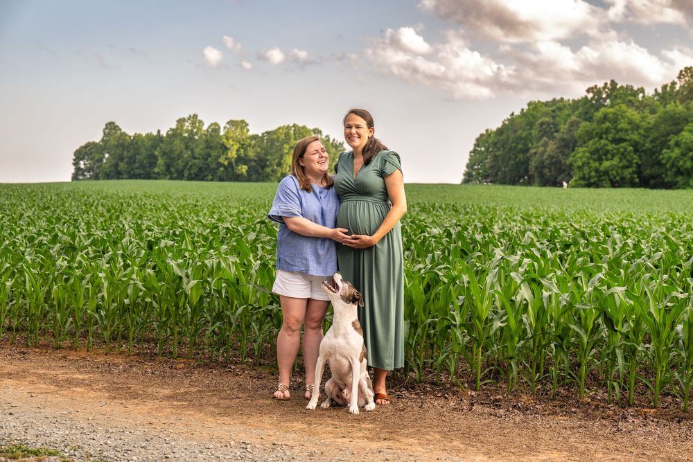 Two people and a dog stand together on a dirt path next to a lush green cornfield on a summer evening.