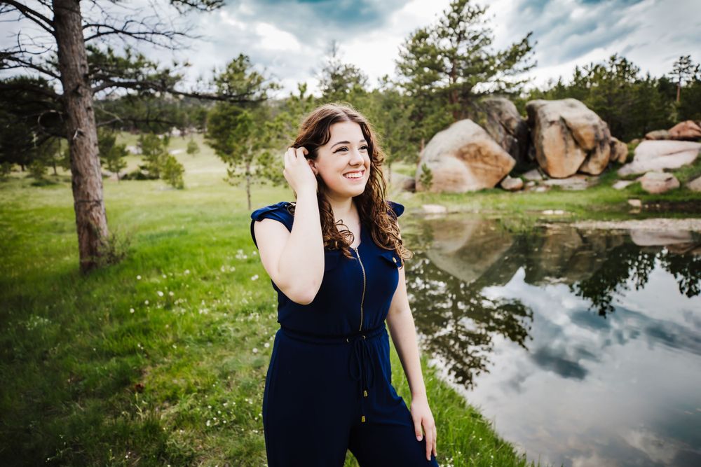 Senior girl in black jumper smiling in grassy field with trees and water near her in Dickson, TN