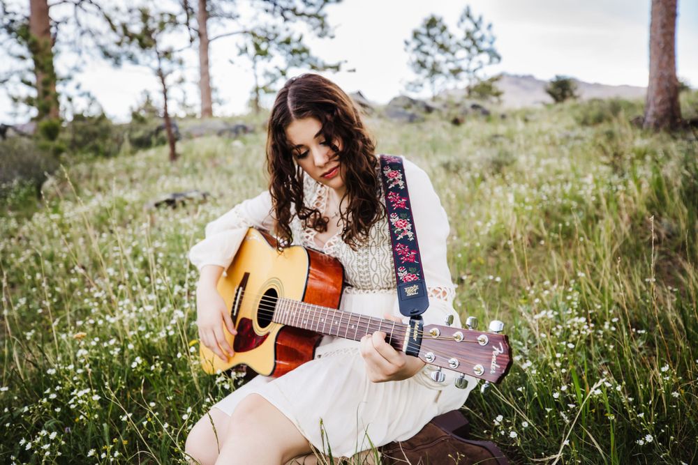 Senior girl in white dress playing her guitar while sitting in flower covered field in Dickson, TN