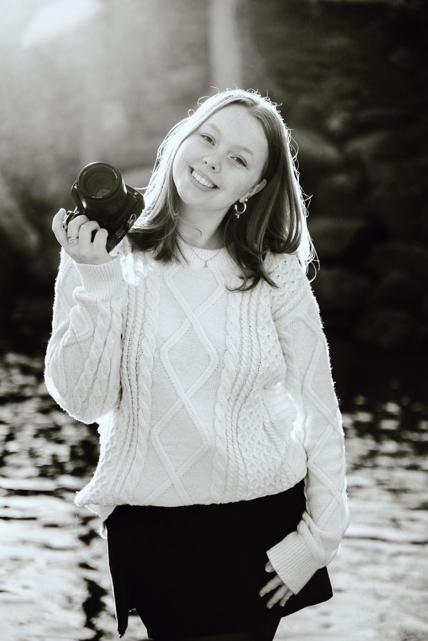 A photographer in a cozy white sweater holds a camera while smiling outdoors in warm backlit sunshine.