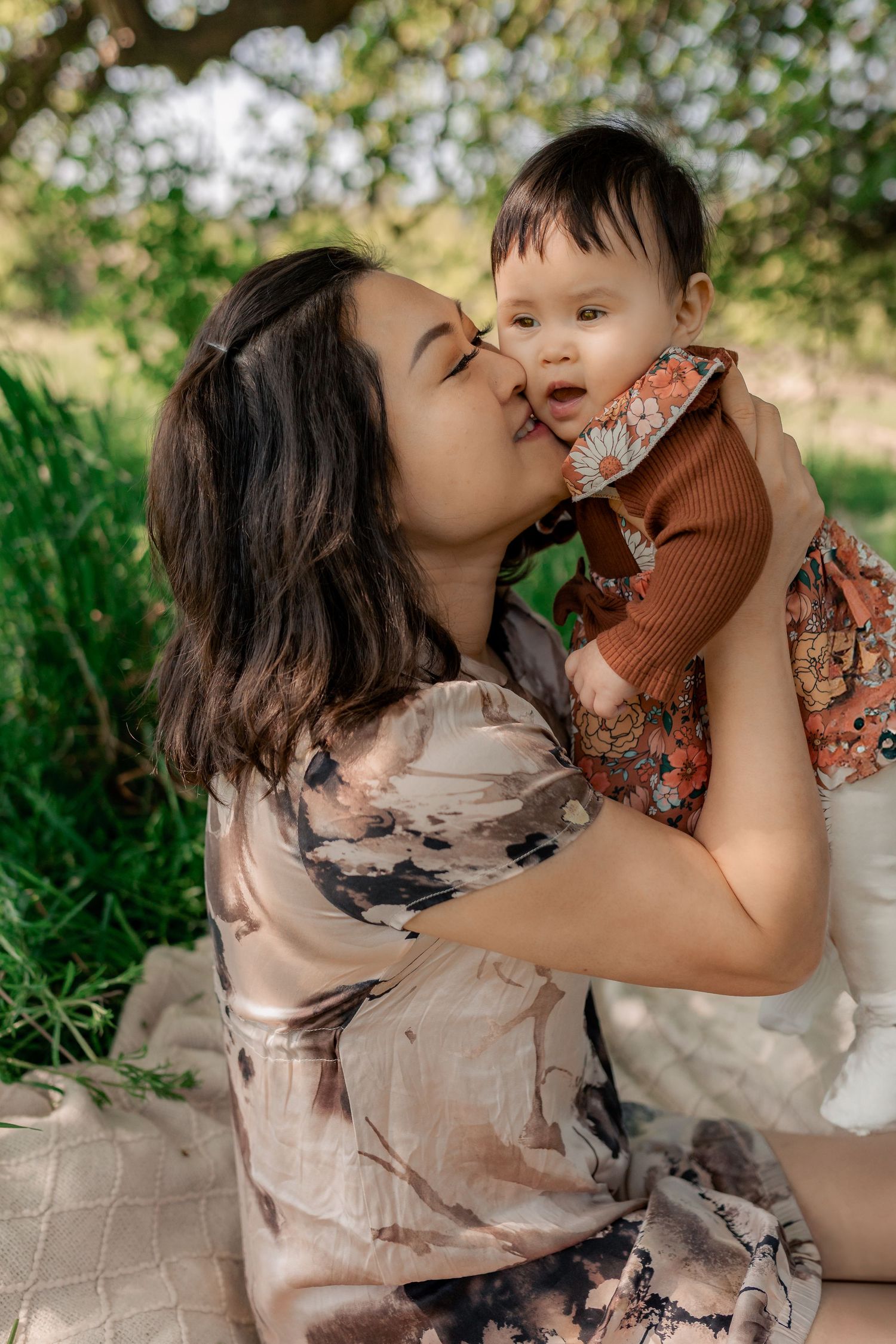 a mom sitting on a blanket beneath a tree while her daughter hugs her from behind.
