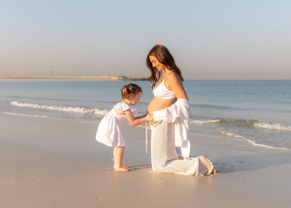Pregnant mother and young daughter on the beach in Dubai at sunrise with daughter holding mum's big belly during maternity photoshoot