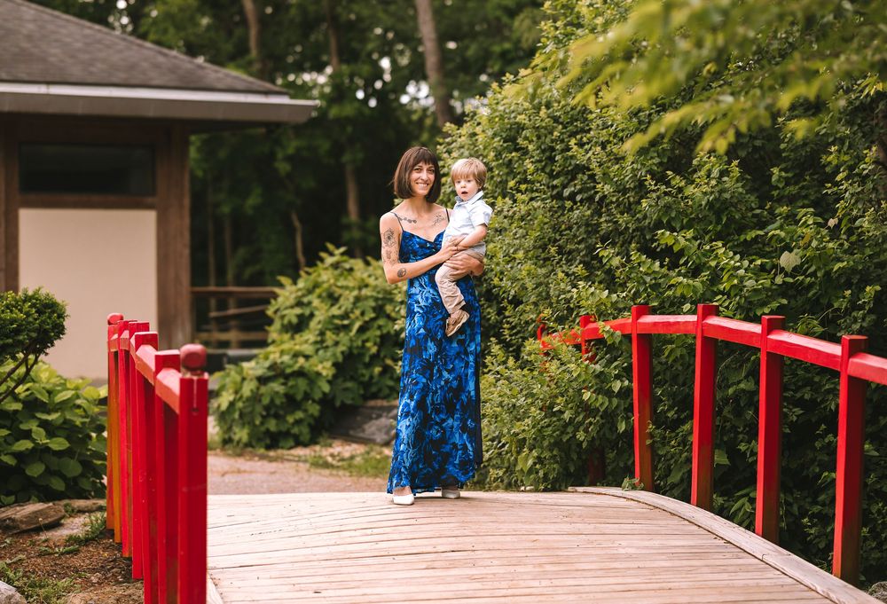 A figure wearing a blue floral dress stands on a red wooden footbridge surrounded by lush greenery.