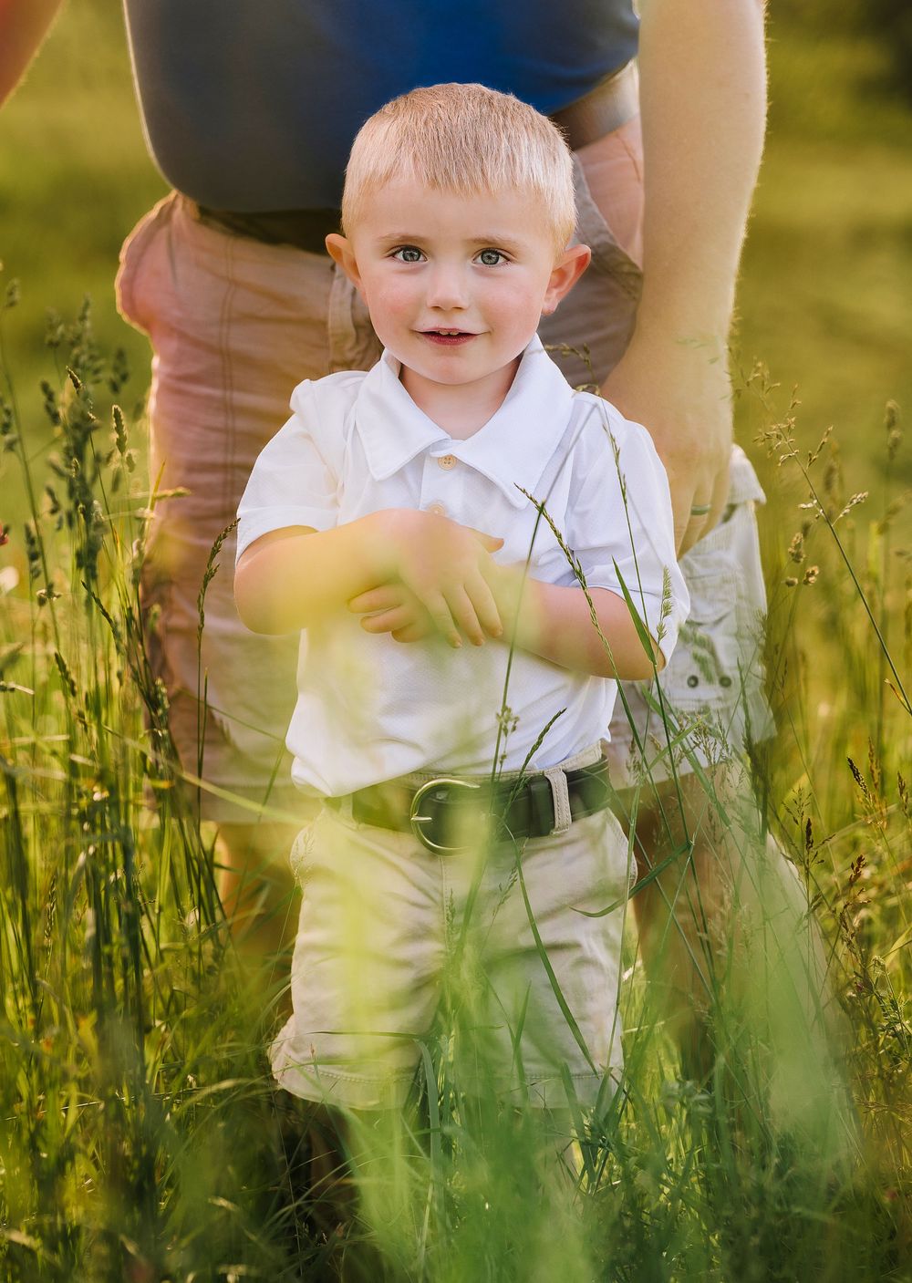 A small child in white clothing stands in a field of tall summer grass during golden hour lighting.